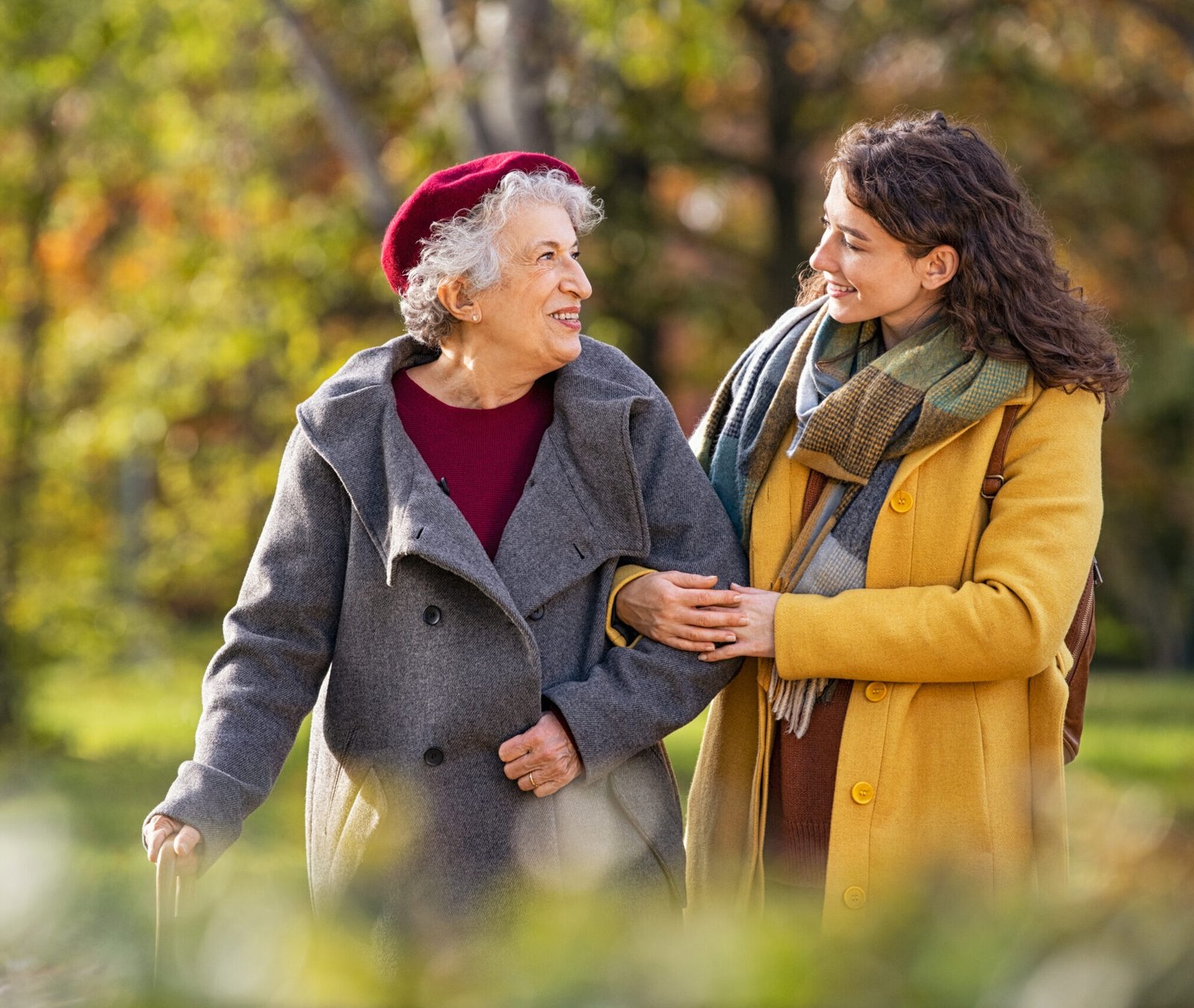 Young woman in park wearing winter clothing walking with old grandmother. Happy grandma wearing coat walking with lovely girl outdoor with copy space. Smiling lovely caregiver and senior lady walking in park during autumn and looking at each other.