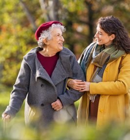 Young woman in park wearing winter clothing walking with old grandmother. Happy grandma wearing coat walking with lovely girl outdoor with copy space. Smiling lovely caregiver and senior lady walking in park during autumn and looking at each other.
